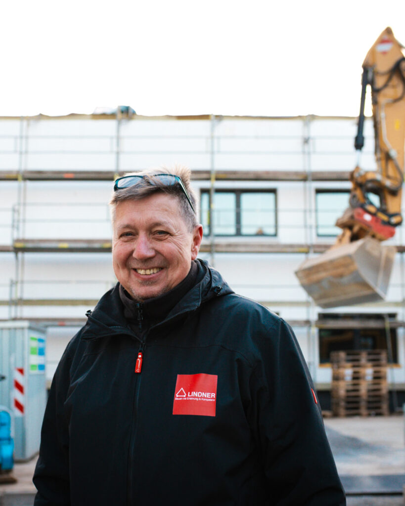 Porträt eines lächelnden Mitarbeiters von Lindner Bau in Firmenjacke vor einem Bauprojekt mit Bagger im Hintergrund.