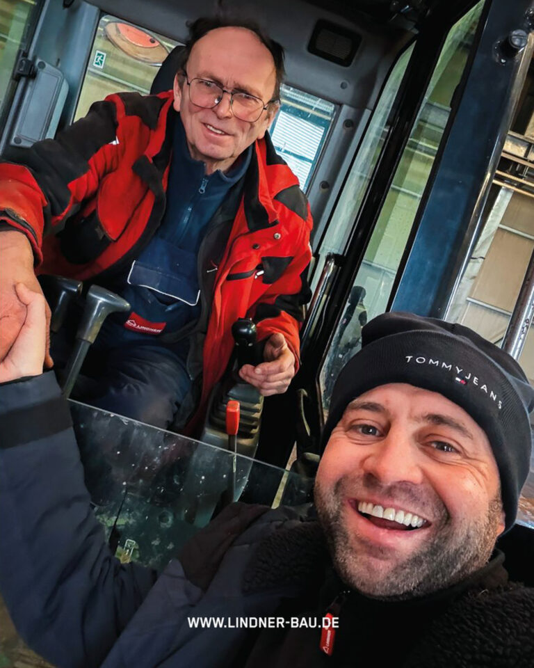Zwei lächelnde Bauarbeiter der Lindner Bau GmbH in der Kabine eines Liebherr Baggers.