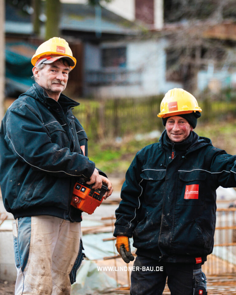 Zwei lächelnde Bauarbeiter der Firma Lindner Bau in Arbeitskleidung und gelben Schutzhelmen auf einer Baustelle.