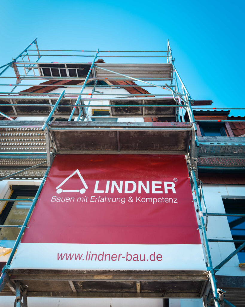 Nahaufnahme eines roten Lindner Bau Banners an einem verzinkten Baugerüst vor einem Wohnhaus.