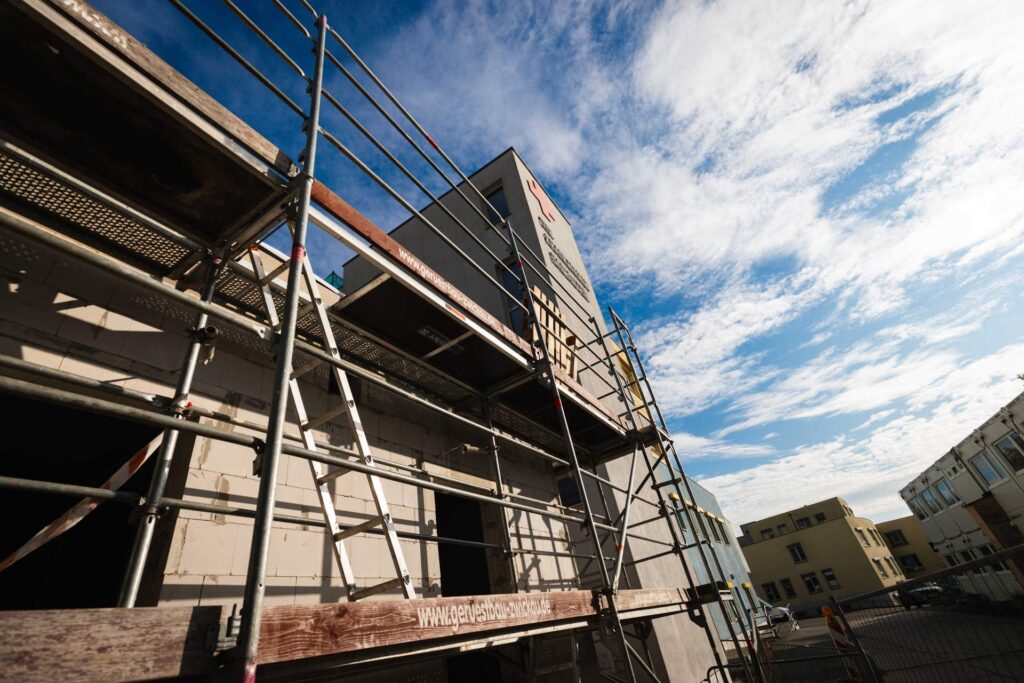 Außenansicht der Fassade mit Baugerüst und Blick auf die neu konstruierte Attika am Dachrand bei blauem Himmel.