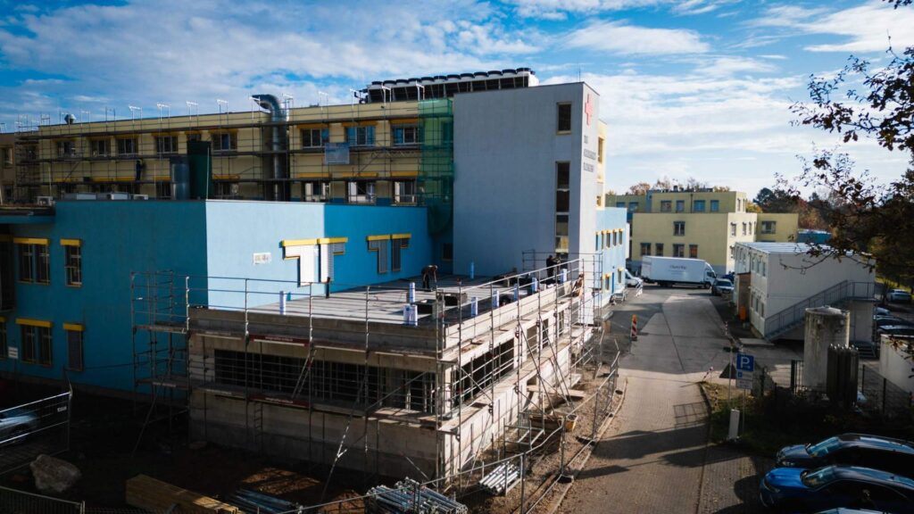 Draufsicht auf die Baustelle des Flachdachanbaus mit sichtbarem Mauerwerk, Gerüstsystemen und Anschluss an das bestehende blaue Klinikgebäude.