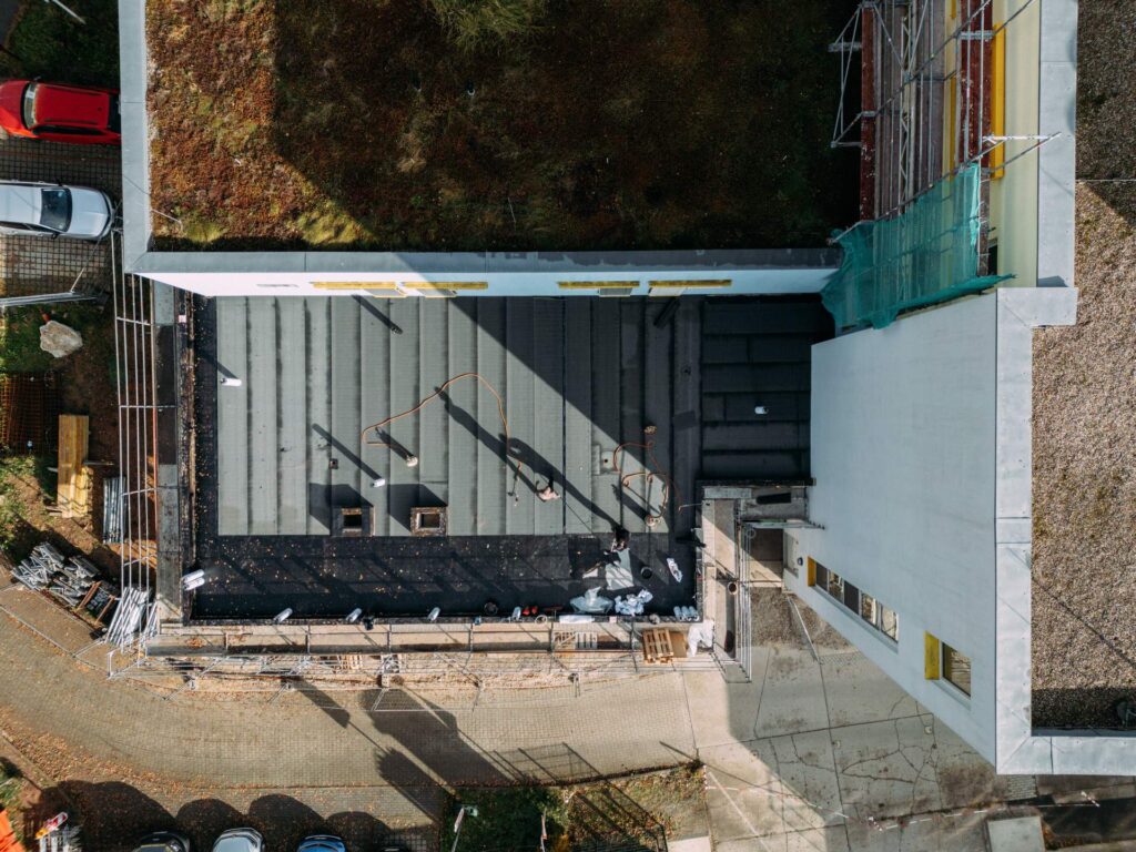 Draufsicht: Vogelperspektive auf das flache Dach mit frisch verlegten Bitumenbahnen, der umlaufenden Attika und dem Kontrast zum bestehenden Gründach.