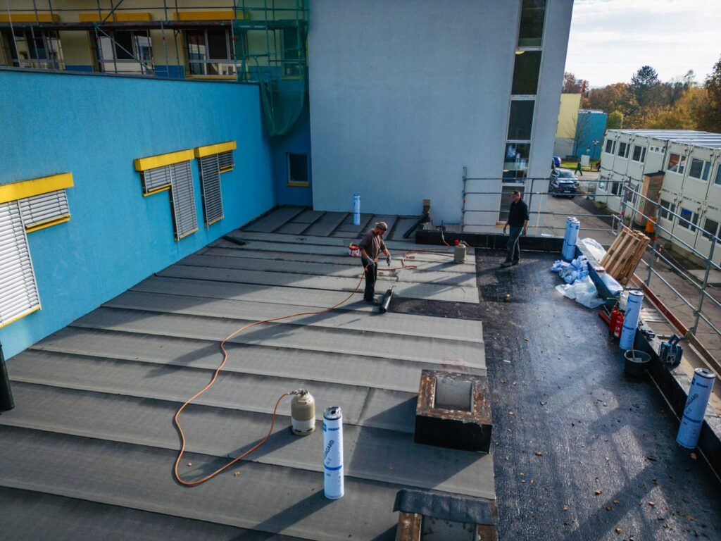 Letzte handwerkerarbeiten auf der Attika: Dachdecker bei der Flachdachabdichtung mit Bitumenschweißbahnen im Bereich der Attika und Wandanschlüsse eines modernen Erweiterungsbaus.