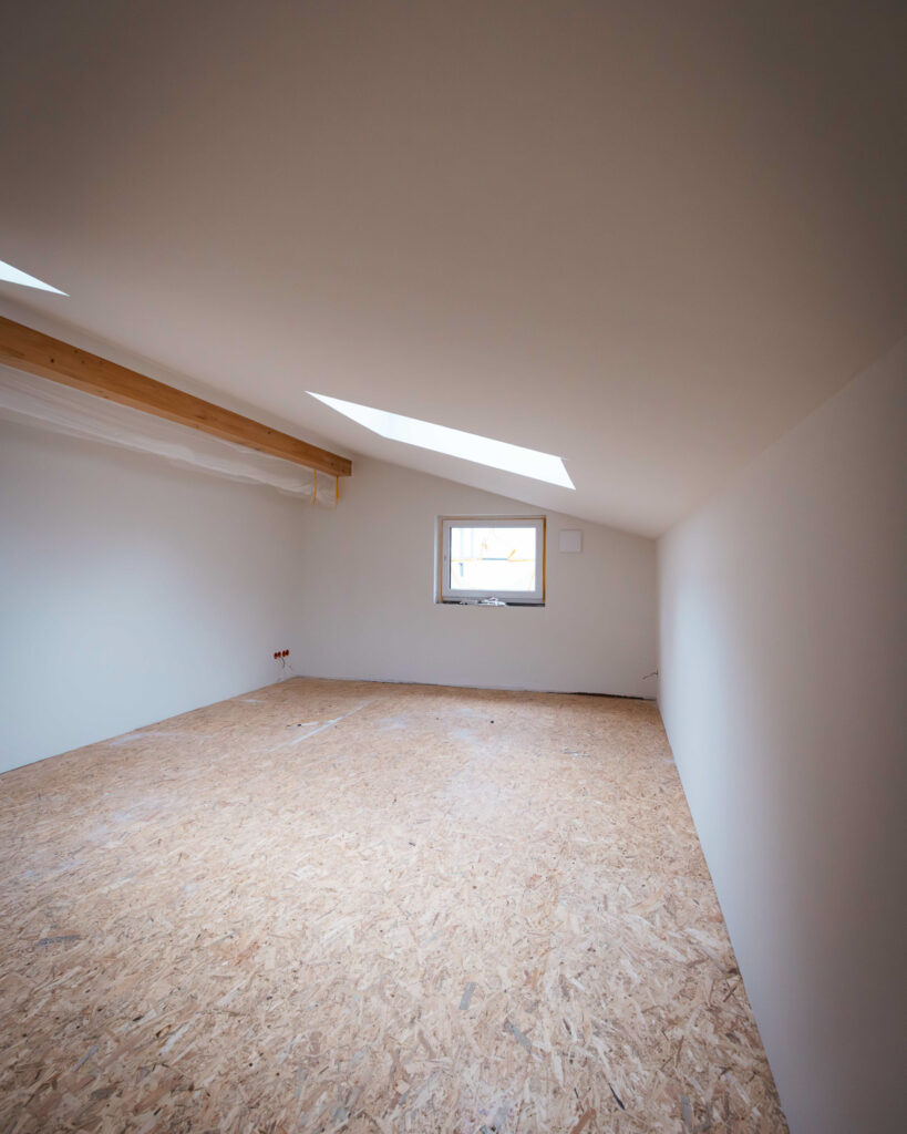 Blick in einen langen Raum unter der Dachschräge mit sichtbarem Holzbalken und Fenster.