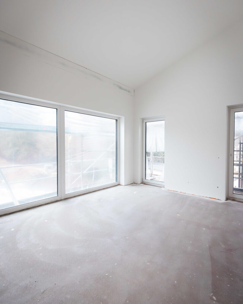 Lichtdurchfluteter Raum im Rohbau mit großen Fensterfronten und frisch verspachtelten Wänden.