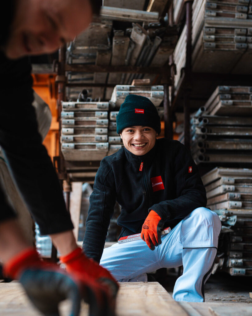 Lächelnder junger Auszubildender im Bauhandwerk kniet mit Zollstock vor gelagerten Schalungselementen.