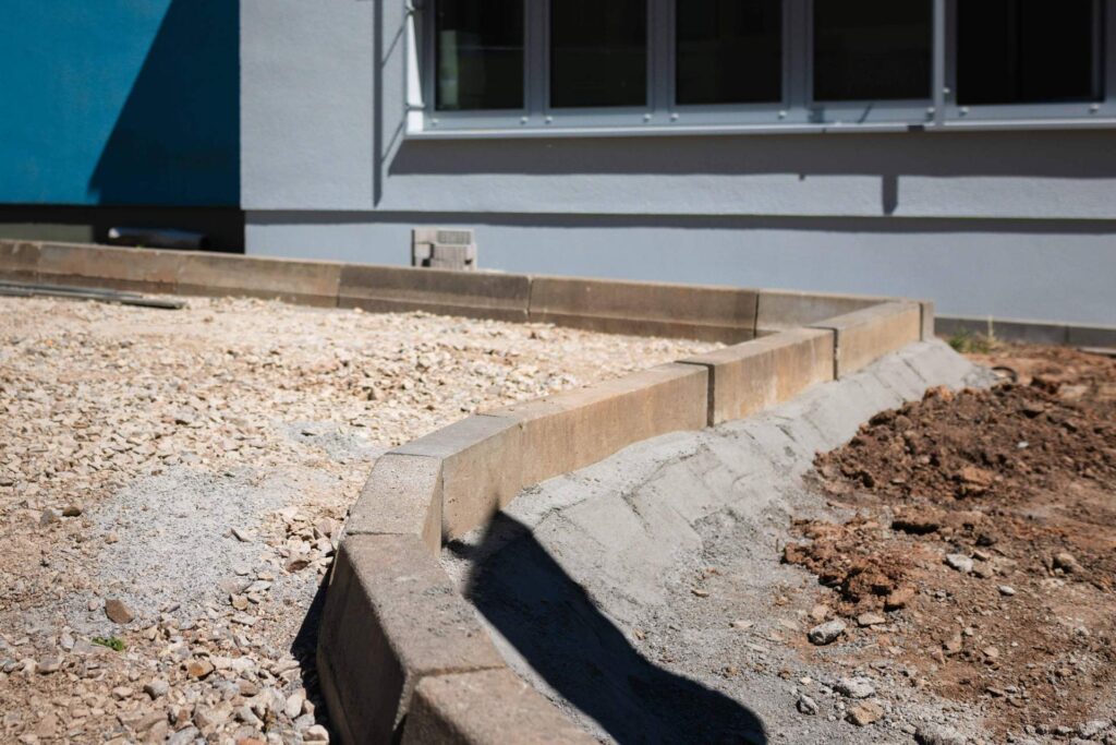 Fachgerechtes Setzen von Bordsteinen in Betonfundament zur Einfassung von Gehwegen und Grünflächen auf dem Krankenhausgelände.