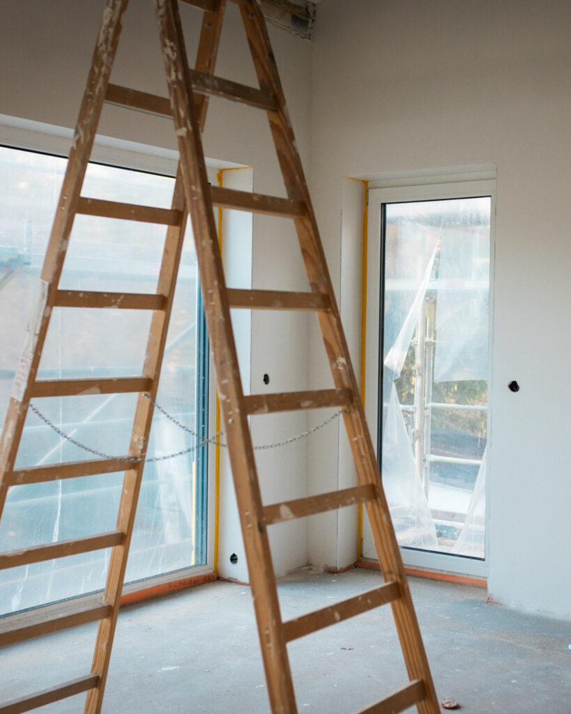 Innenraum auf der Baustelle mit einer hölzernen Doppel-Stehleiter vor großflächigen, bodentiefen Fenstern.