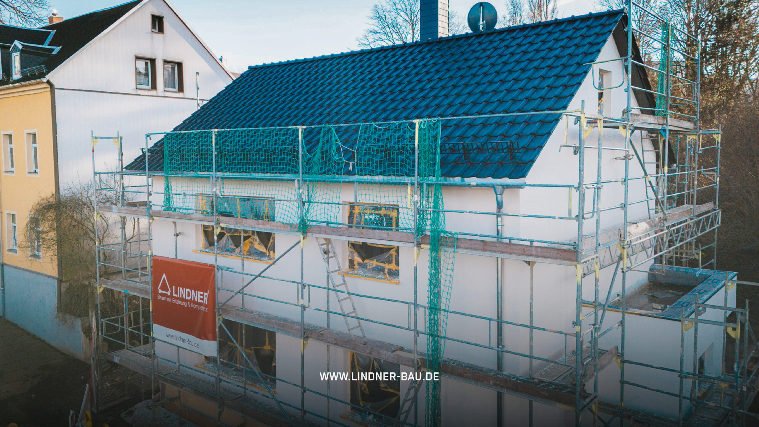 Einfamilienhaus im Rohbau mit weiß verputzter Fassade und blauem Ziegeldach, eingerüstet mit einem Baugerüst der Firma Lindner Bau.