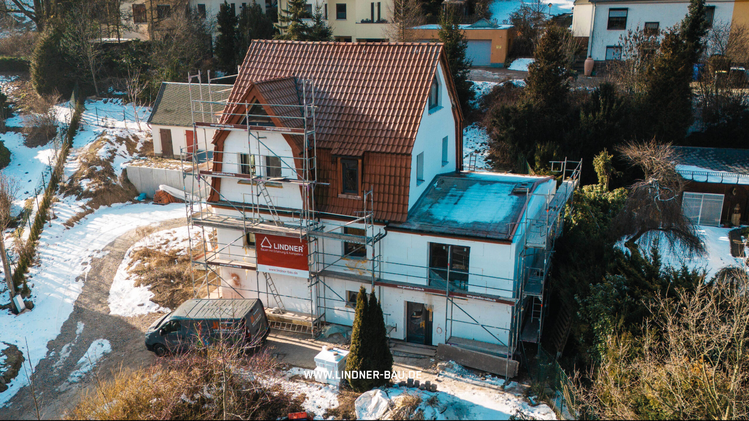 Luftaufnahme eines Einfamilienhauses während der Fassadensanierung mit Gerüstbau, Lindner Bau Werbebanner und Firmenfahrzeug in winterlicher Umgebung.