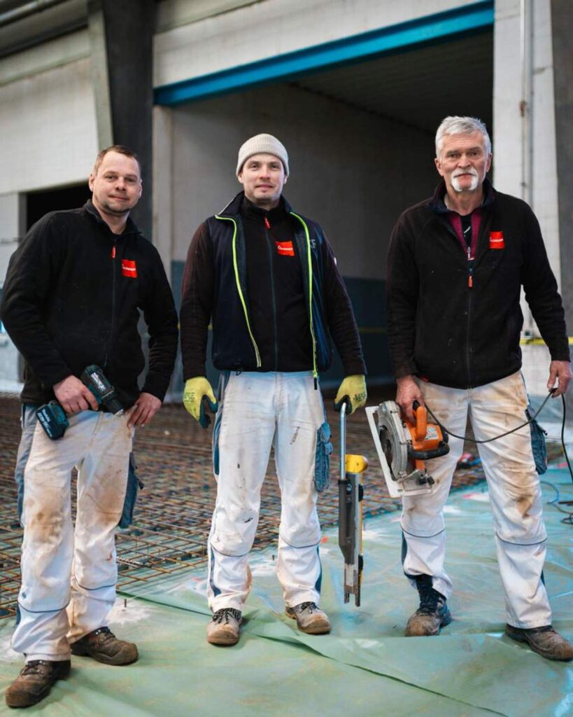 Drei Facharbeiter von Lindner Bau posieren mit Werkzeugen (Akkuschrauber, Kreissäge) auf der Bewehrung in der Industriehalle Chemnitz.