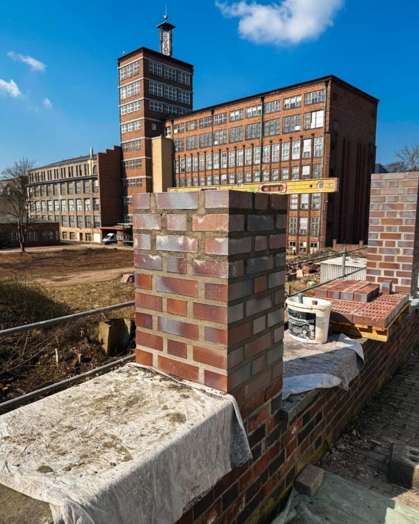 Detaillierte Nahaufnahme eines neu errichteten Ziegelpfeilers, der auf einer Mauerkrone steht. Im Hintergrund erstreckt sich die große, historische rote Backsteinfabrik mit ihrem Uhrturm unter blauem Himmel.