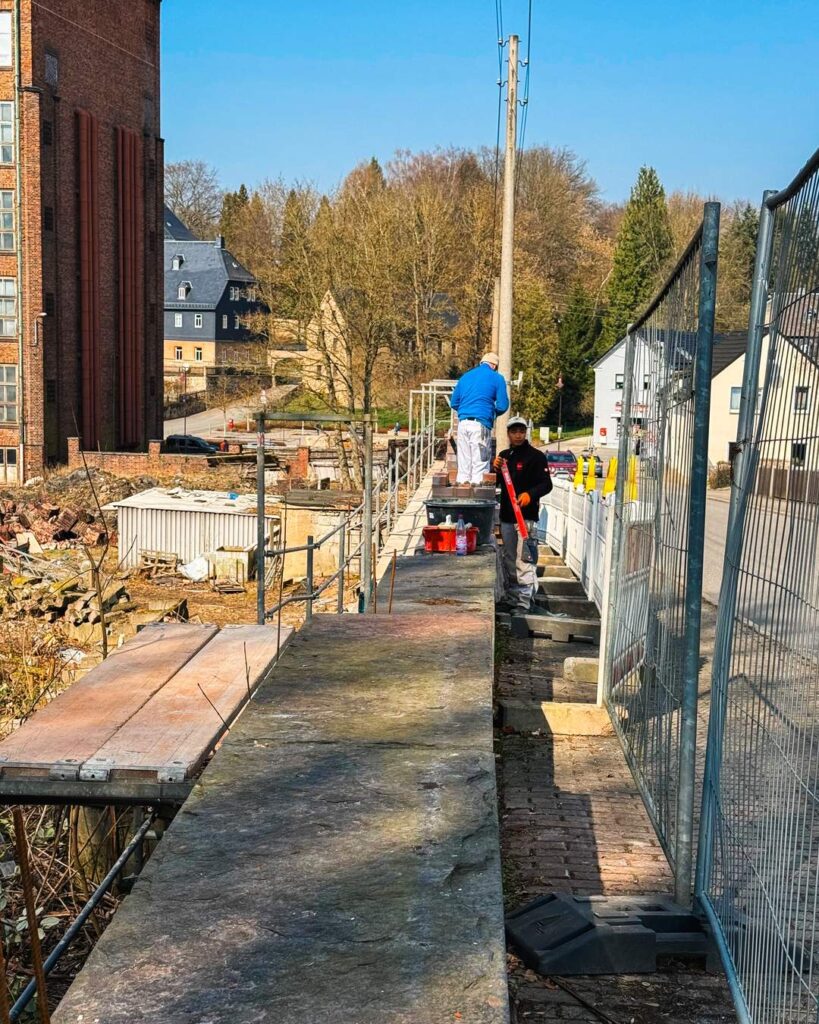 Zwei Bauarbeiter in Schutzkleidung arbeiten auf einem schmalen Gerüstgang an einer Mauer. Einer trägt eine blaue Jacke, der andere eine schwarze. Im Hintergrund sind ein Hang mit Bäumen und Wohngebäude zu sehen.