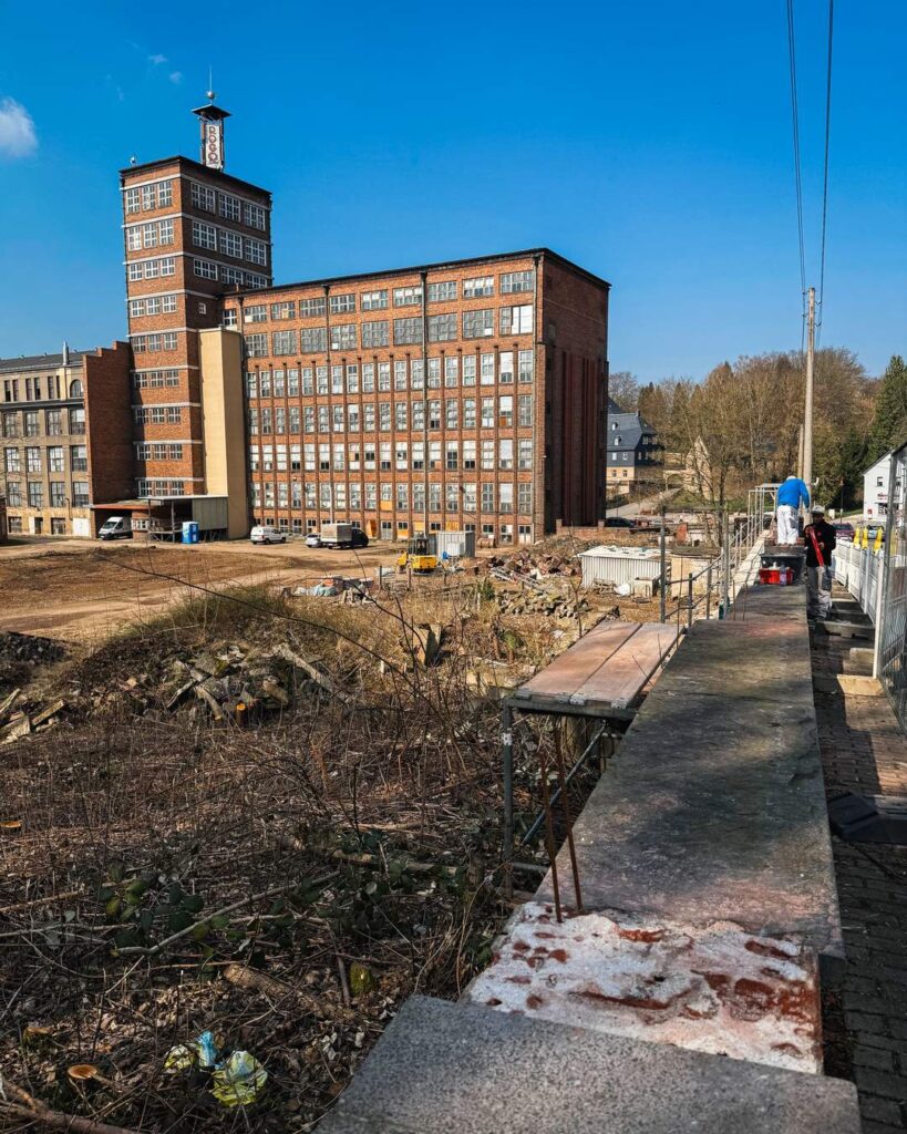 Blick von einer erhöhten Betonmauer über ein brachliegendes Baugelände mit Gestrüpp und Baumaterialien auf eine große, historische Backsteinfabrik mit Uhrturm. Zwei Bauarbeiter arbeiten am rechten Rand.