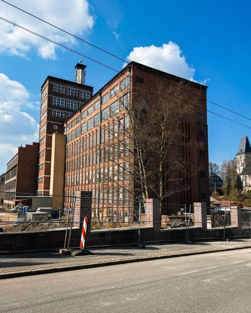 Große, renovierungsbedürftige rote Backsteinfabrik mit einem markanten Uhrturm und vielen Fenstern. Ein Bauzaun im Vordergrund grenzt das Gelände zur Straße ab. Eine kahle Baumgruppe steht vor dem Gebäude. Blauer Himmel mit Wolken.