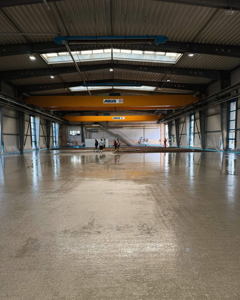 Frisch gegossener, spiegelglatter Betonboden in einer großen Lagerhalle mit gelben ABUS Deckenkranen im Hintergrund.
