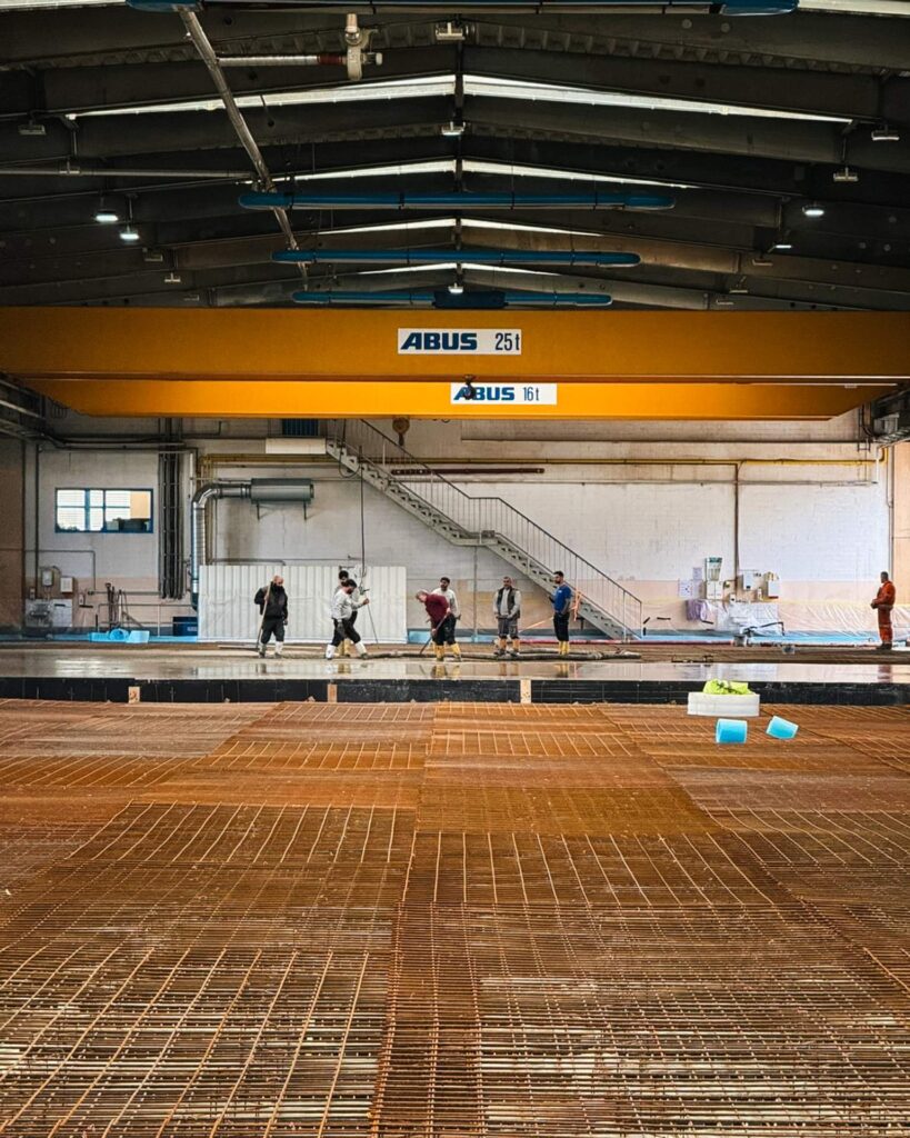 Bauarbeiter-Team beim fachgerechten Verteilen und Nivellieren von Flüssigbeton über einer Stahlbewehrung in einer Werkhalle.