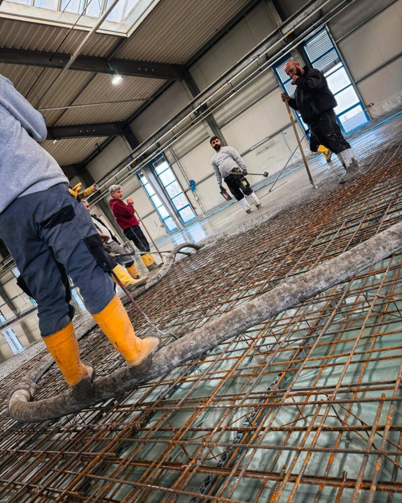 Detailaufnahme der Betonage: Bauarbeiter in gelben Gummistiefeln führen den Betonschlauch über die Bewehrungsmatten.