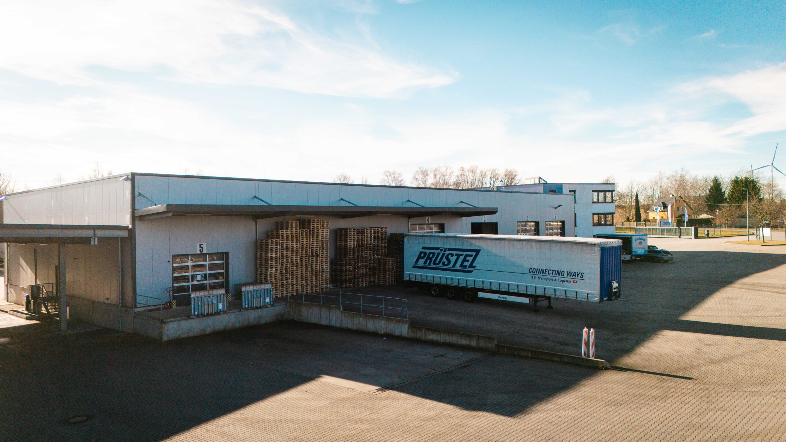 Außenansicht eines Logistikzentrums der Firma Prüstel mit einem Lkw-Auflieger an der Laderampe, gestapelten Holzpaletten und einem modernen Lagergebäude unter blauem Himmel.