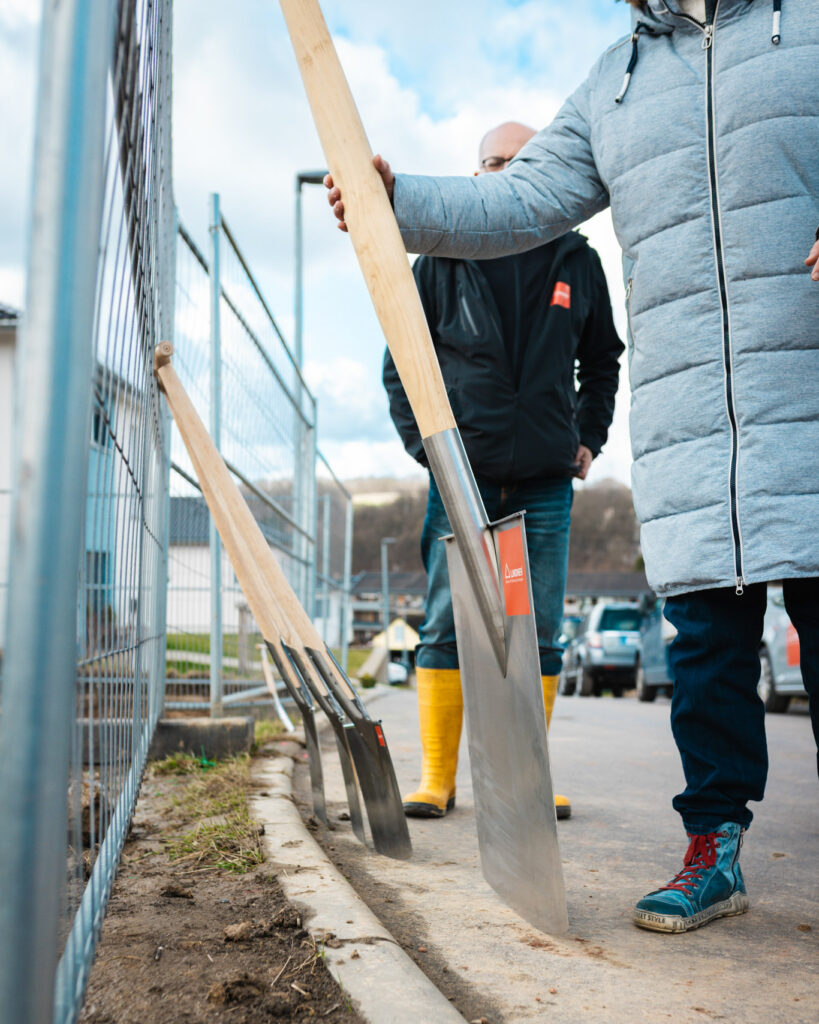 Nahaufnahme von mehreren Spaten, die für den Spatenstich an einer Baustelle bereitstehen, im Hintergrund ein Bauzaun und Mitarbeiter in Arbeitskleidung.
