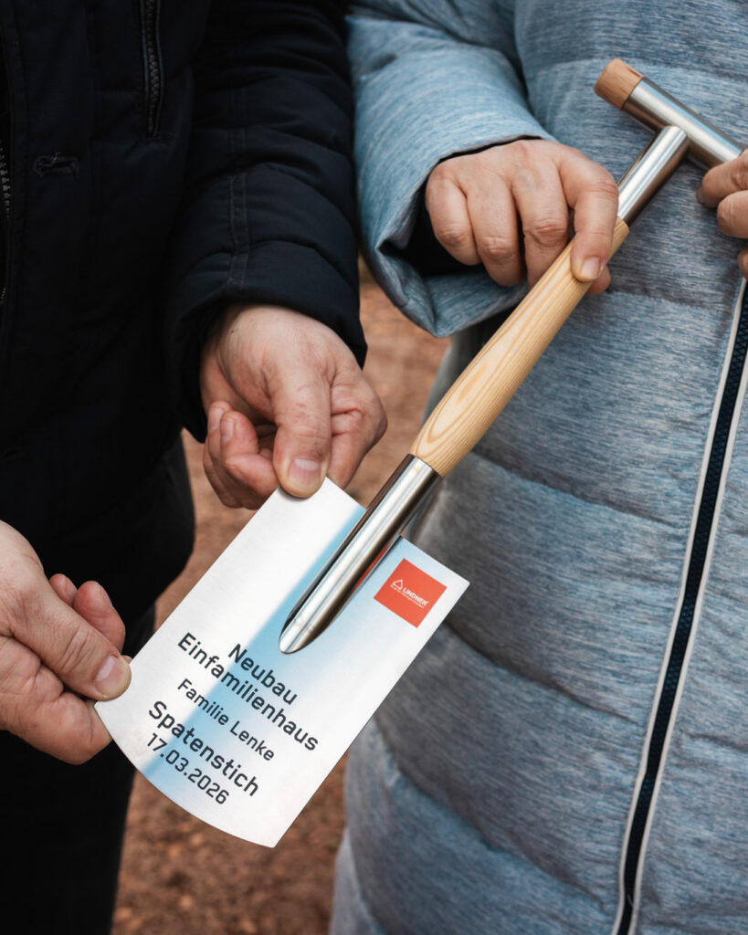 Hände halten einen gravierten Miniatur-Spaten mit der Aufschrift „Neubau Einfamilienhaus Familie Lenke – Spatenstich 17.03.2026“ und dem Lindner-Bau Logo.