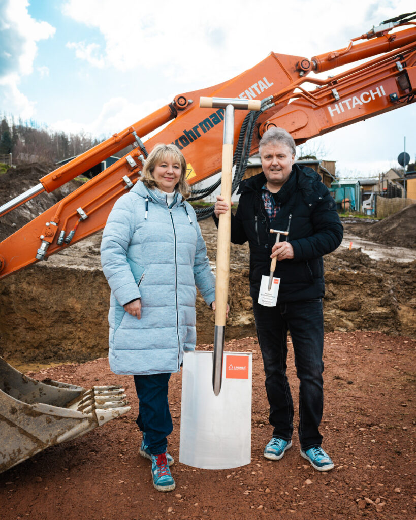 Das Bauherren-Ehepaar Lenke posiert glücklich mit einem großen silbernen Spaten und einem Miniatur-Spaten vor einem Hitachi-Bagger auf ihrem Grundstück.