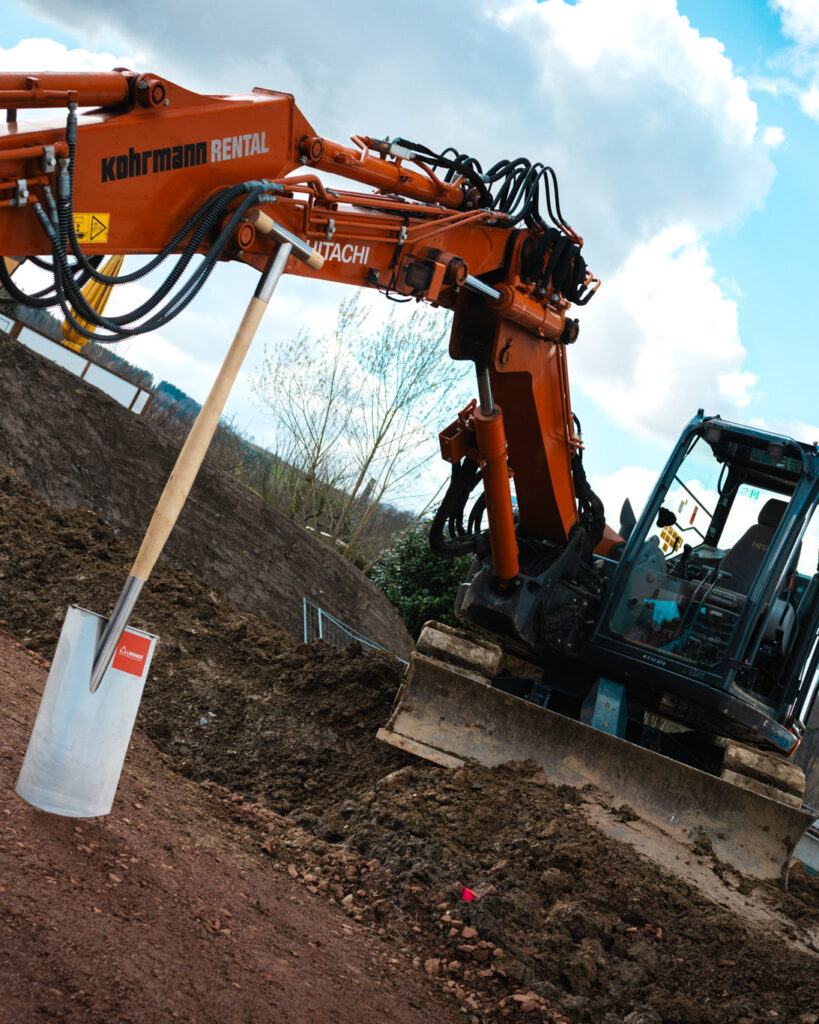 Ein großer orangefarbener Hitachi-Hydraulikbagger auf einer Erdaushub-Baustelle, im Vordergrund steckt ein silberner Spaten von Lindner-Bau in der Erde.