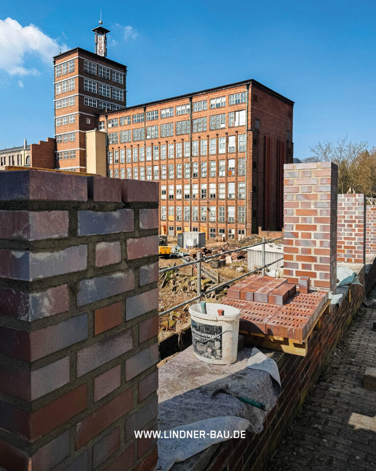 Baustelle mit neuen Ziegelpfeilern im Vordergrund, Blick auf eine historische, mehrstöckige Fabrik aus Backstein mit Uhrturm unter blauem Himmel. Arbeitsmaterialien wie ein Eimer und lose Ziegel sind sichtbar.