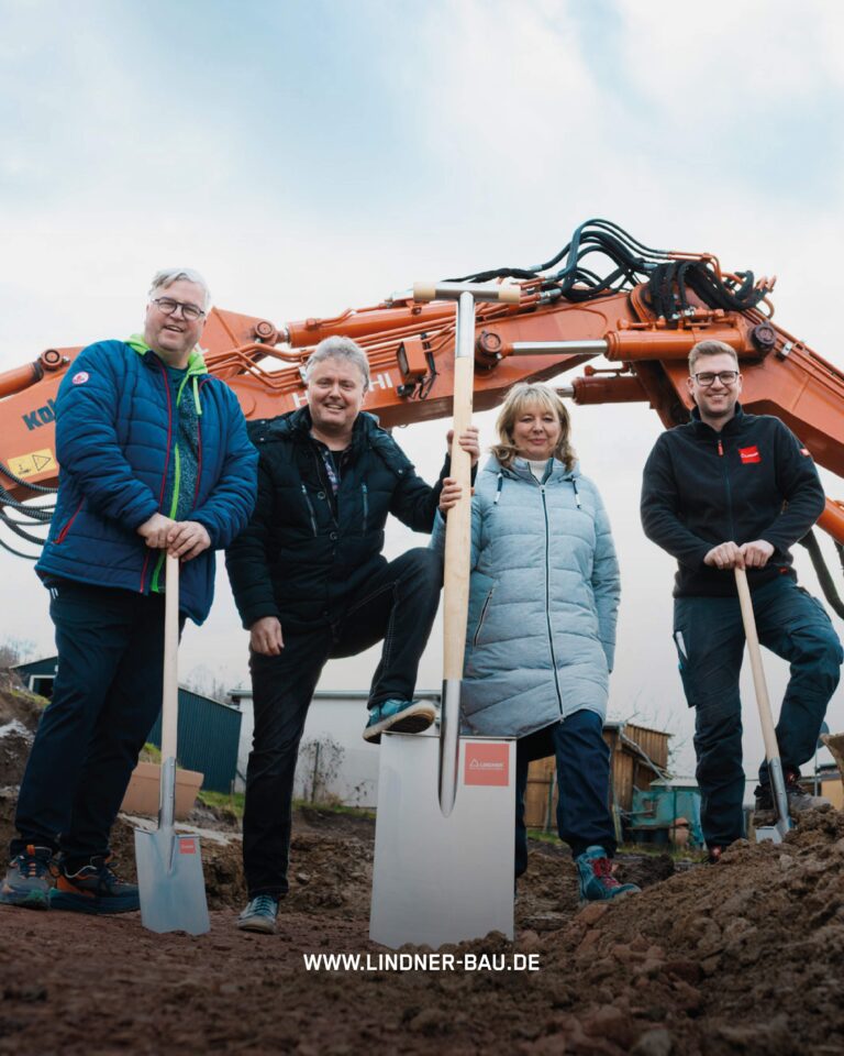 Vier Personen lächeln beim offiziellen Spatenstich für einen Neubau vor einem orangefarbenen Hitachi-Bagger.