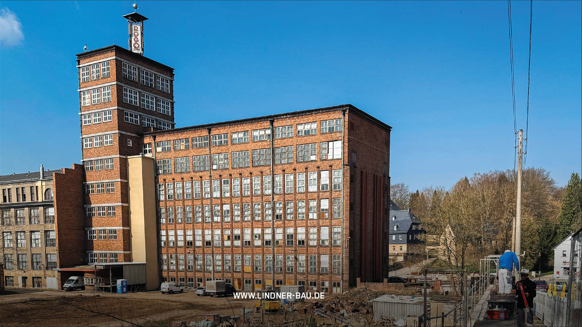 Totale einer imposanten, mehrstöckigen Backsteinfabrik mit Uhrturm und zahlreichen Fenstern. Der Innenhof ist eine unbefestigte Baustelle mit parkenden Fahrzeugen. Zwei Bauarbeiter arbeiten rechts auf einem Gerüst.