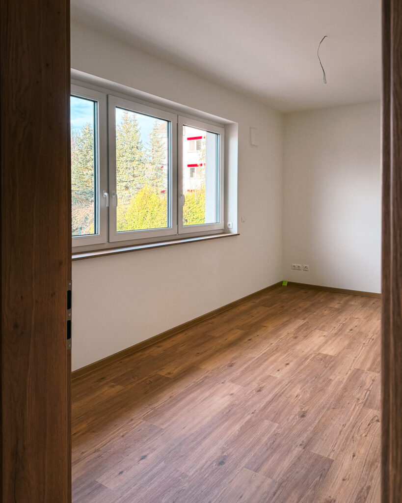 Heller Wohnraum mit großem Fenster, Holzfußboden und weiß gestrichenen Wänden im bezugsfertigen Neubau.