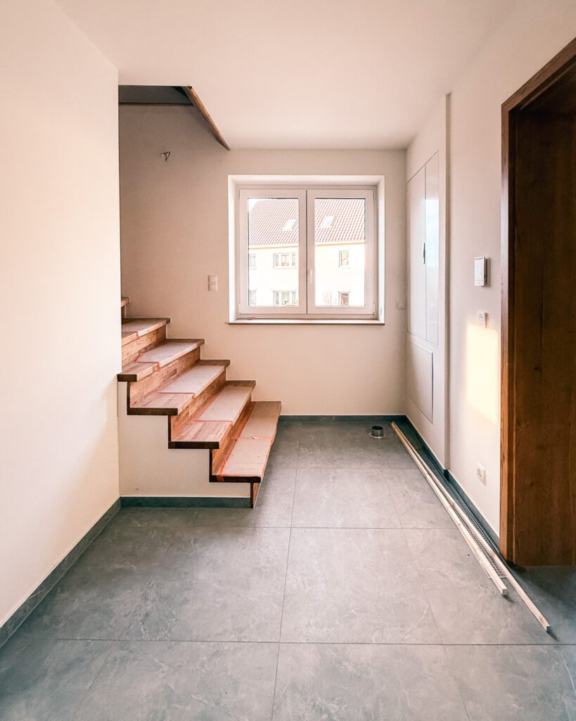 Moderner Eingangsbereich mit großformatigen grauen Fliesen und einer Holztreppe, die in das Obergeschoss führt.