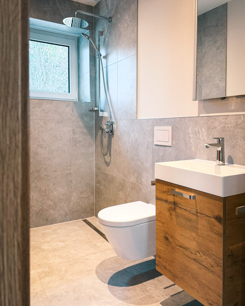 Modernes Badezimmer mit bodengleicher Dusche, Regenduschkopf, hellgrauen Fliesen und Waschtisch mit Holzunterschrank.