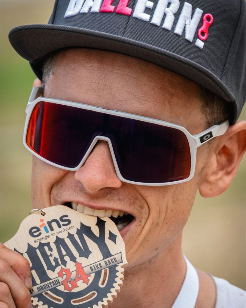 Nahaufnahme eines Radrennfahrers mit Sport-Sonnenbrille, der stolz auf seiner hölzernen Heavy24 Finisher-Medaille beißt