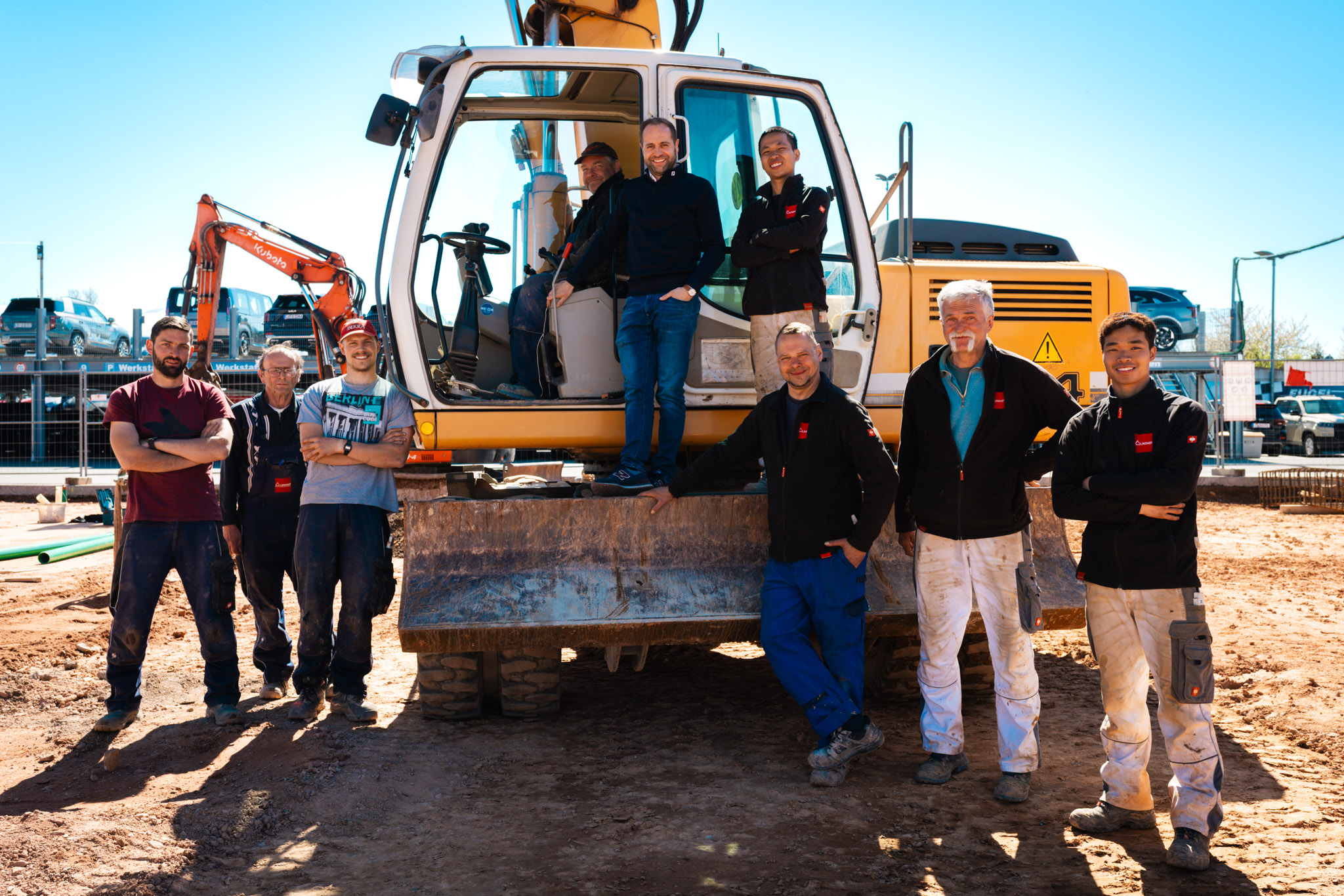 das Team auf der baustelle des Autohauses Fugel in mittelbach posiert mit einem Bagger vor der sonnigen Baustelle.