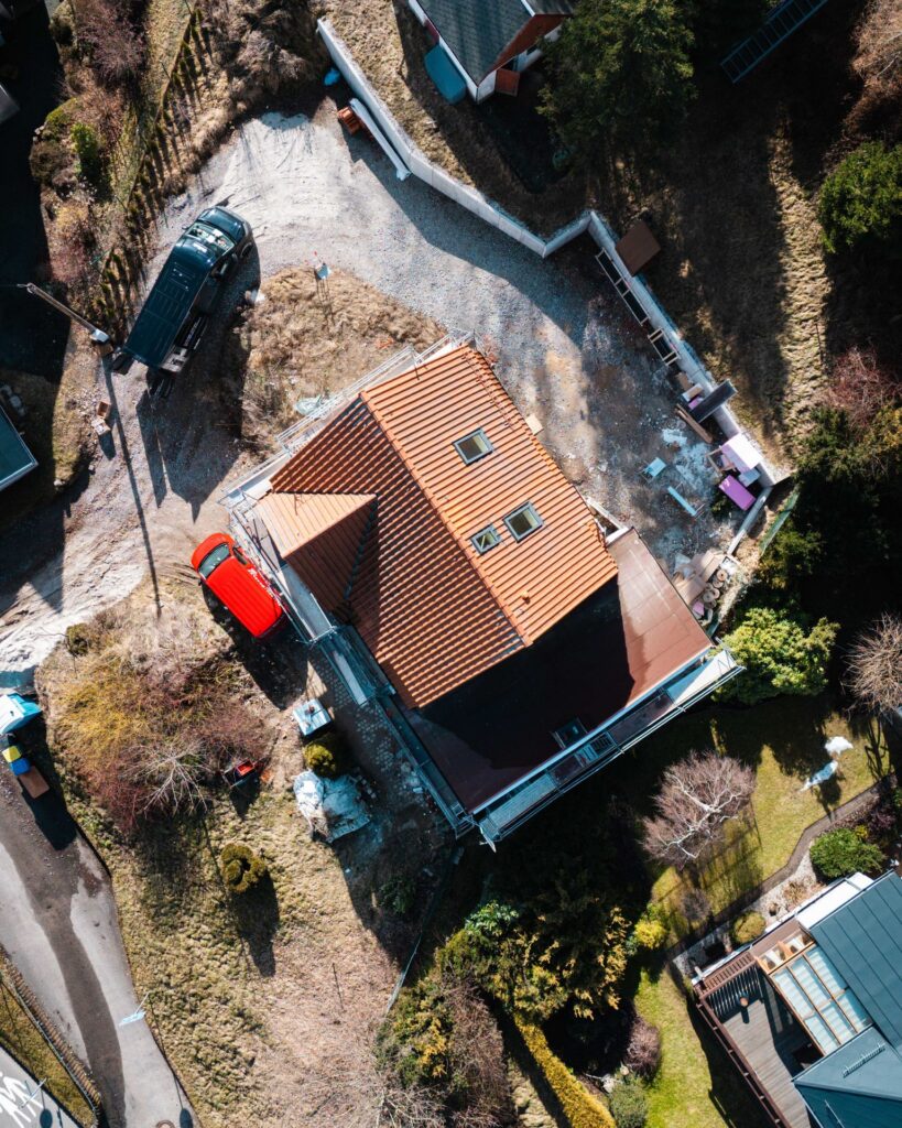 Luftaufnahme einer Baustelle mit Einfamilienhaus, rotem Ziegeldach, Baugerüst und Handwerker-Transportern.