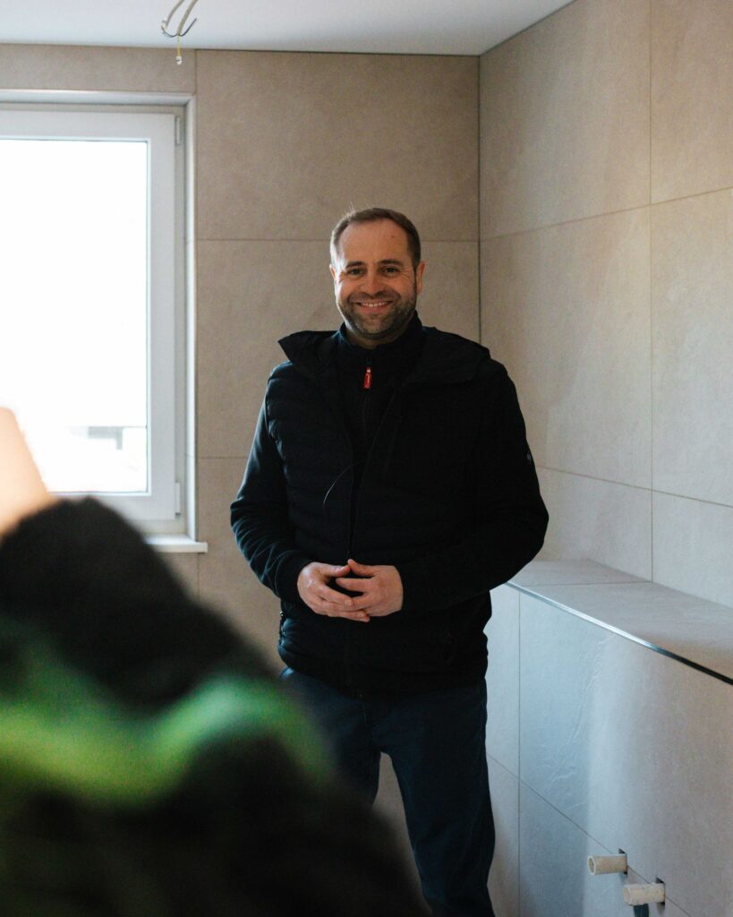 Lächelnder Bauleiter in Arbeitskleidung bei der Begehung eines neu gefliesten Badezimmers im Neubau.