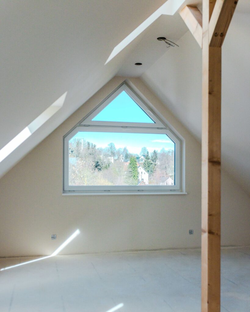 Helles Dachgeschoss im Rohbau mit großem, dreieckigem Giebelfenster und Blick ins Grüne.