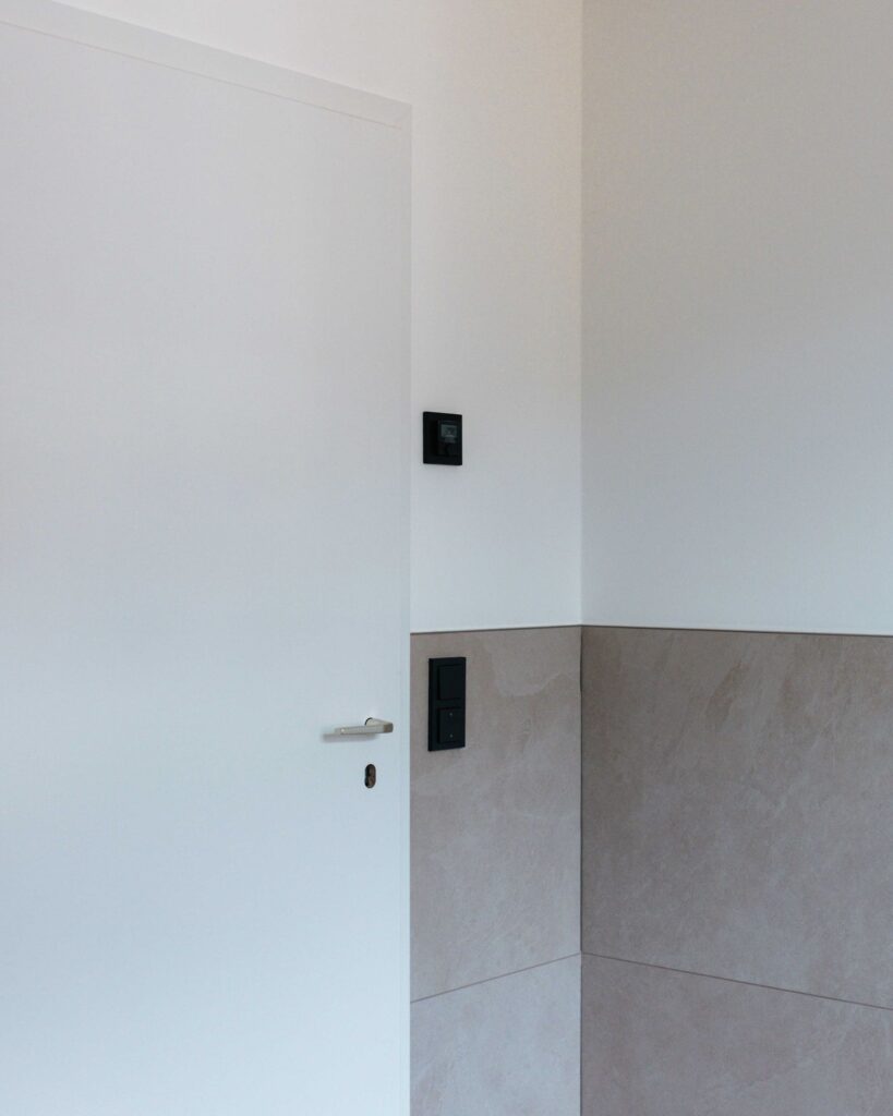 Moderne weiße Zimmertür im Neubau neben mattschwarzen Lichtschaltern und beigen Wandfliesen.