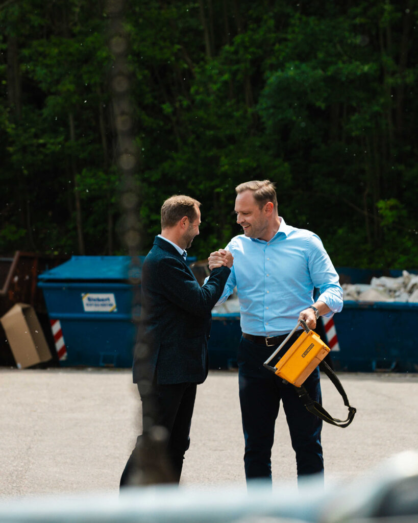 Herzliche Begrüßung auf dem Bauhof: Tony Lindner und Landtagspräsident Alexander Dierks besiegeln das Treffen mit einem festen, lächelnden Handschlag im Außenbereich. Im Hintergrund sind große blaue Container und Bäume zu sehen.