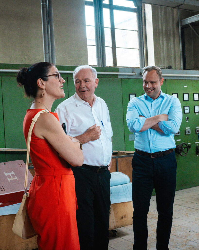 Diskussion vor historischer Kulisse: Ina Klemm im Gespräch mit Thomas Hetzel und Alexander Dierks, der mit verschränkten Armen und einem Lächeln zuhört. Im Hintergrund befindet sich eine grüne, historische Schalttafel bzw. Industrieanlage.