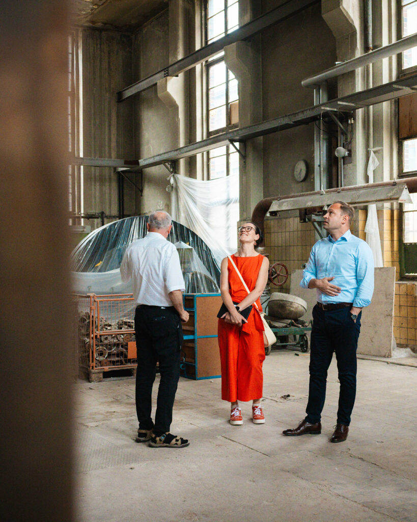 Außenbegehung der ROGO-Werke: Alexander Dierks erklärt gestikulierend ein Detail, während der Oberlungwitzer Bürgermeister Thomas Hetzel, Lars Kluge und Ina Klemm aufmerksam zuhören. Im Hintergrund ist die alte Fabrikfassade zu sehen.