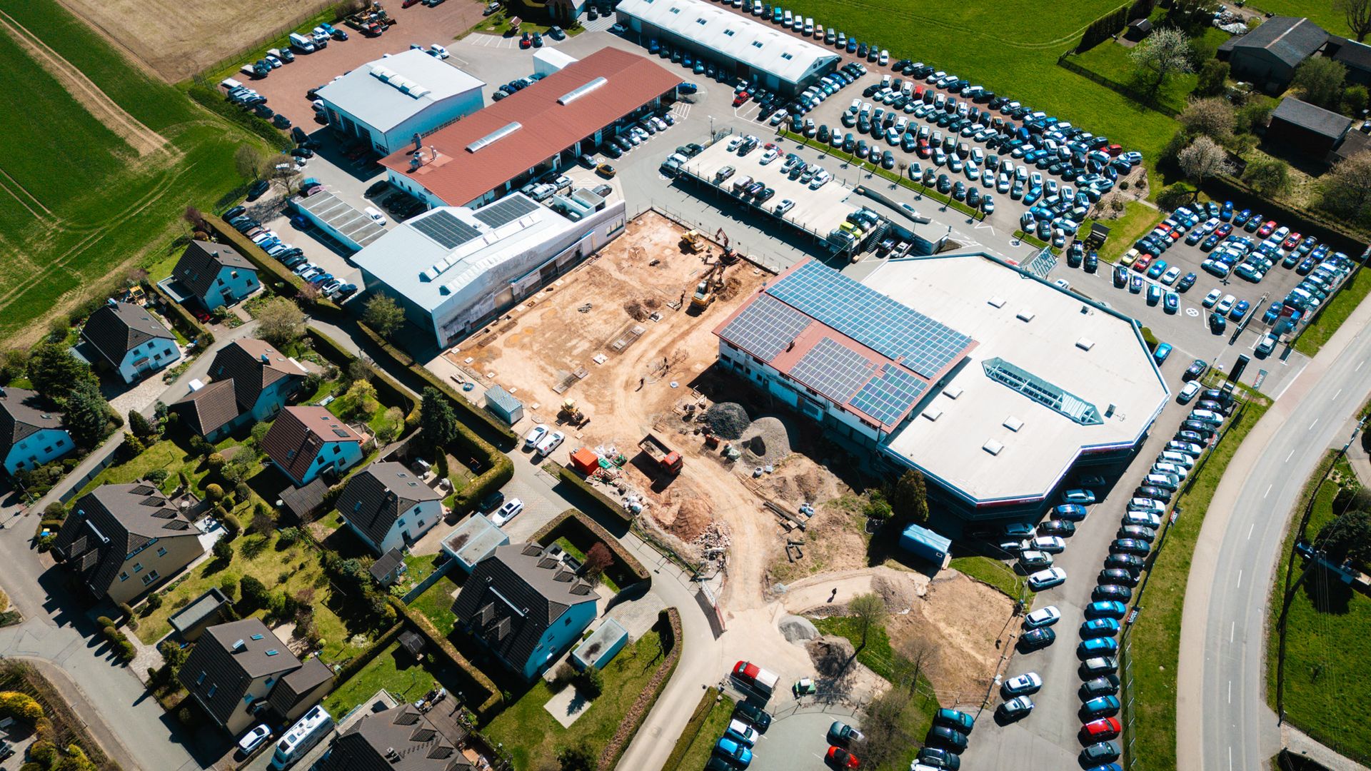 Luftbild eines Autohauses in Deutschland mit einer Baustelle für die Erweiterung. Zu sehen sind Neuwagen, Gebrauchtwagen, Werkstattgebäude, Solarmodule auf einem Dach und angrenzende Wohnbebauung.