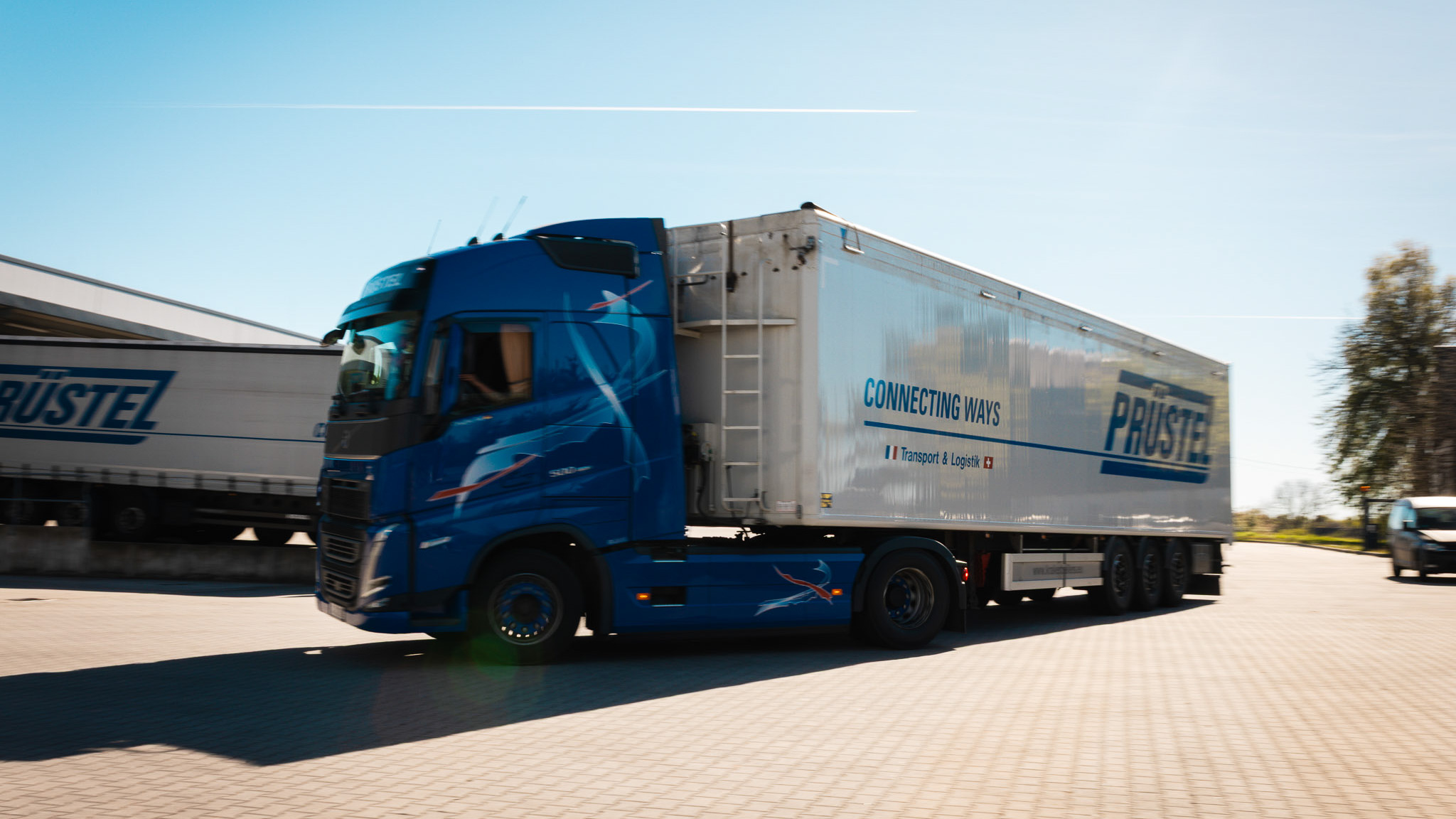 Blauer LKW mit weißem Auflieger von der Spedition Prüstel fährt auf gepflasterter Straße.