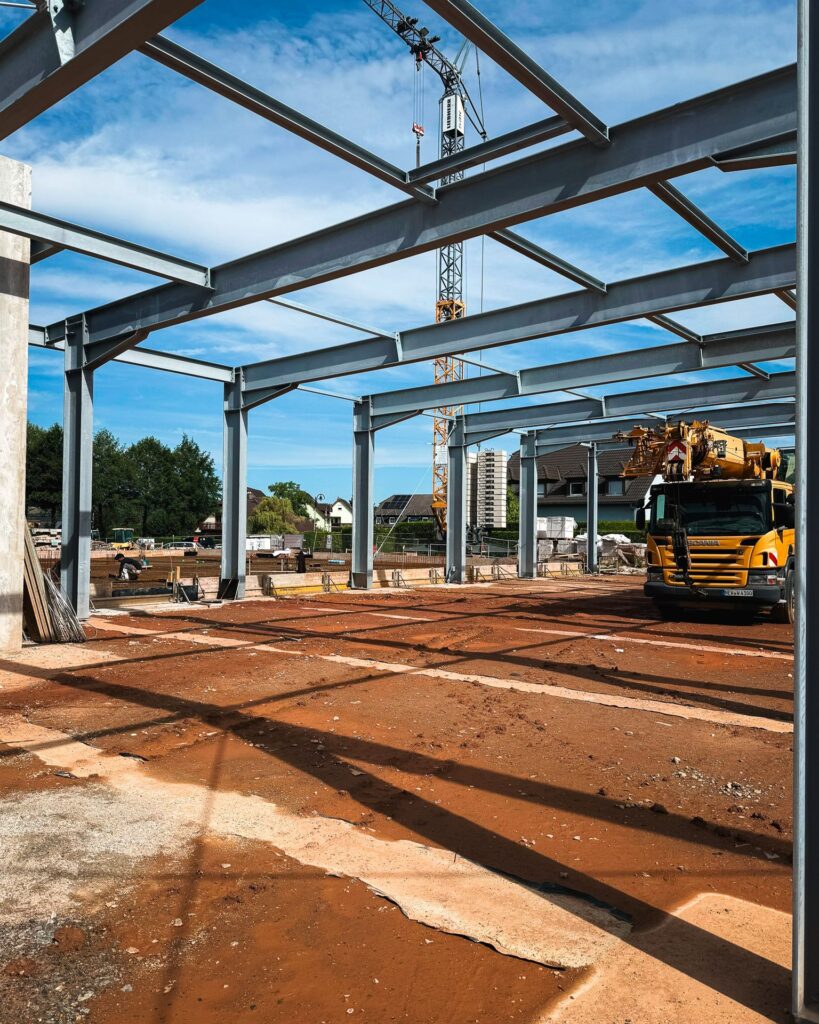 Baustelle für eine Industriehalle: Blick durch das offene Stahltragwerk auf den unbefestigten Boden und einen gelben Autokran im Hintergrund bei blauem Himmel.