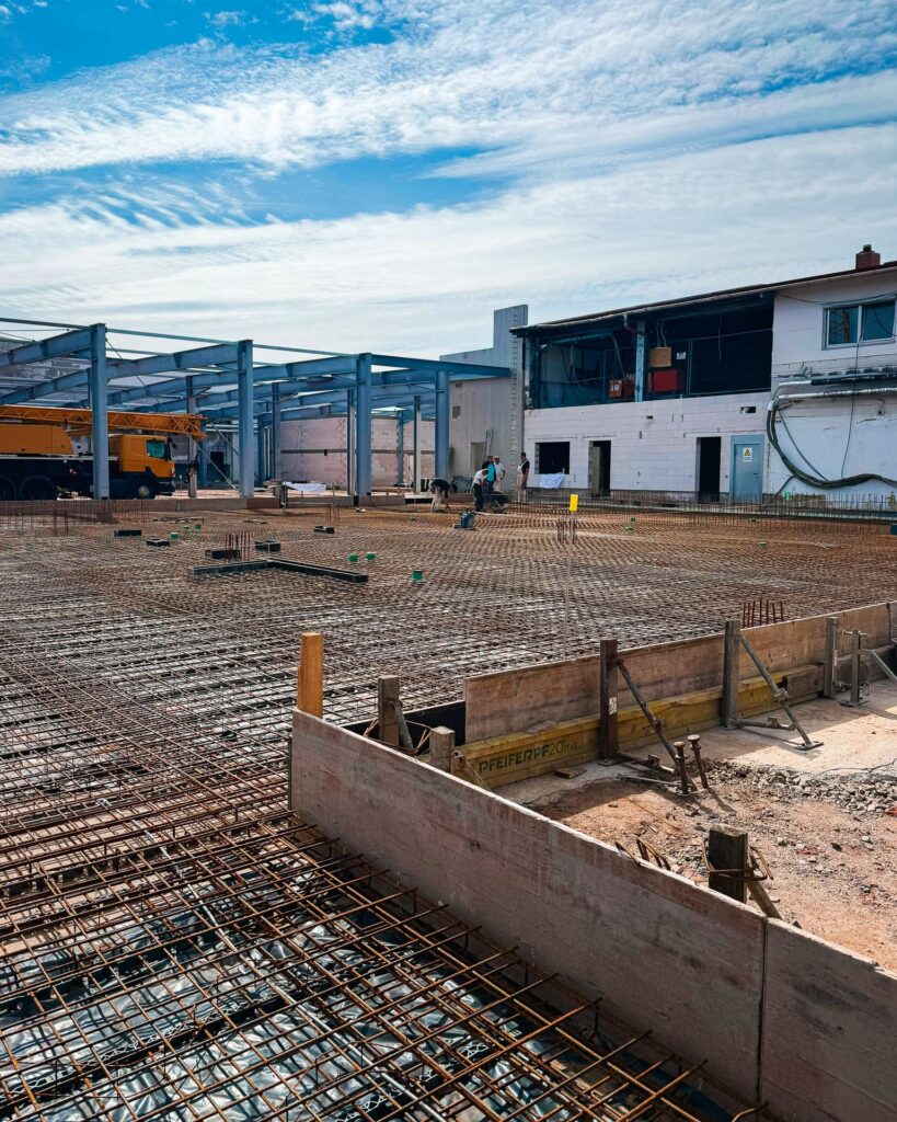 Bewehrung für Bodenplatte: Vorbereitung der Stahlarmierung auf einer Baustelle für einen Hallenanbau, im Hintergrund Arbeiter und ein gelber Kran.