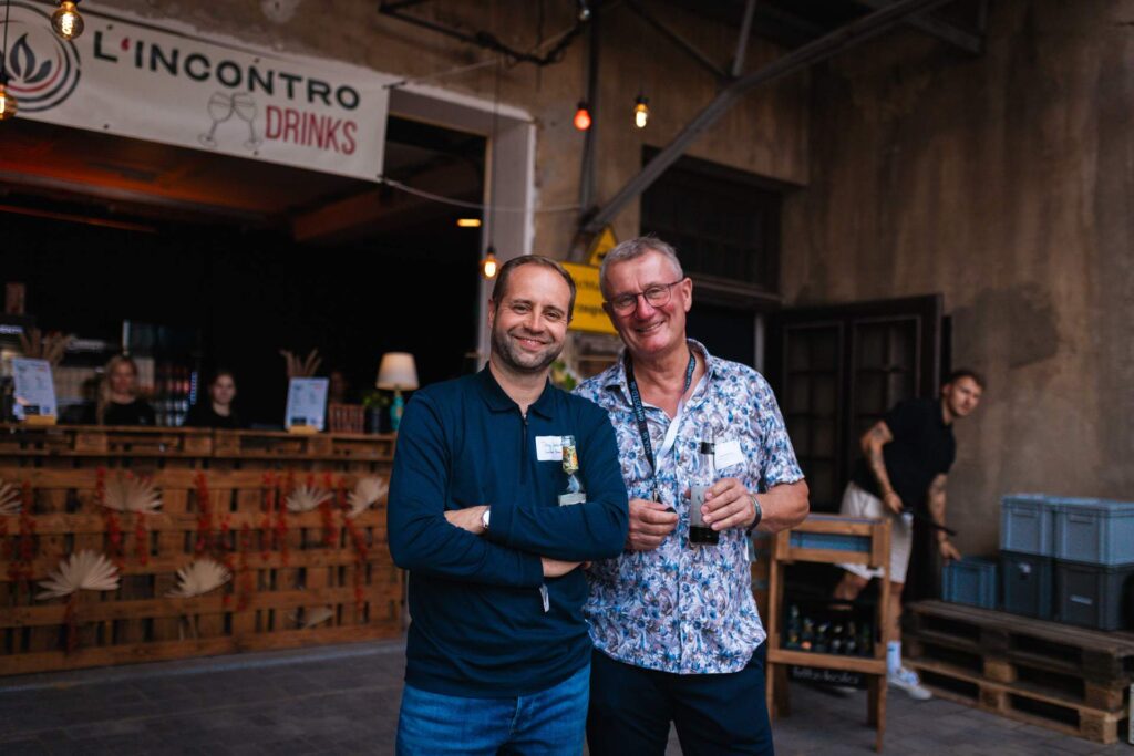 Tony Lindner von Bau-und Transportgesellschaft LINDNER GmbH und ein weiterer Mann, lächeln vor dem Getränkestand von L'Incontro beim Sommerfest des Mittelstands.
