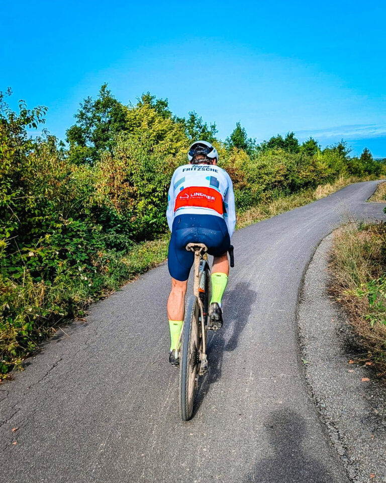 Rückansicht eines Radfahrers auf einem Gravelbike auf einem asphaltierten Weg. Der Fahrer trägt einen weißen Helm, ein weißes Trikot mit blauer „TEAM FRITZSCHE“- und roter „LINDNER“-Aufschrift, dunkelblaue Hosen und neongelbe Socken. Das Rad und die Beine sind leicht mit Schmutz bedeckt. Der Weg führt durch eine grüne Naturlandschaft mit Büschen und Bäumen unter blauem Himmel.