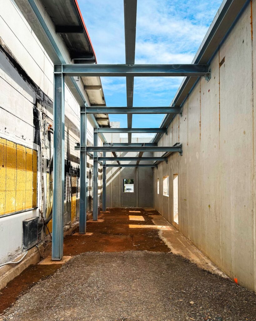 Gang im Hallenrohbau: Perspektivischer Blick durch eine Reihe von Stahlstützen und -trägern, flankiert von einer alten Wand und einer neuen Betonwand.